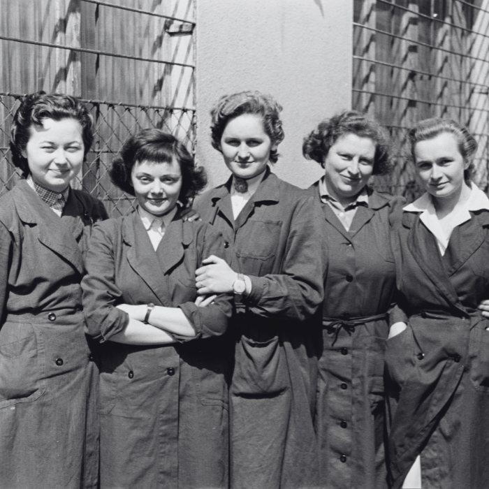 Öt nő áll egy csoportban, mindannyian ugyanabban az egyenruhában, mosolyognak, háttérben egy fémkerítés látható.